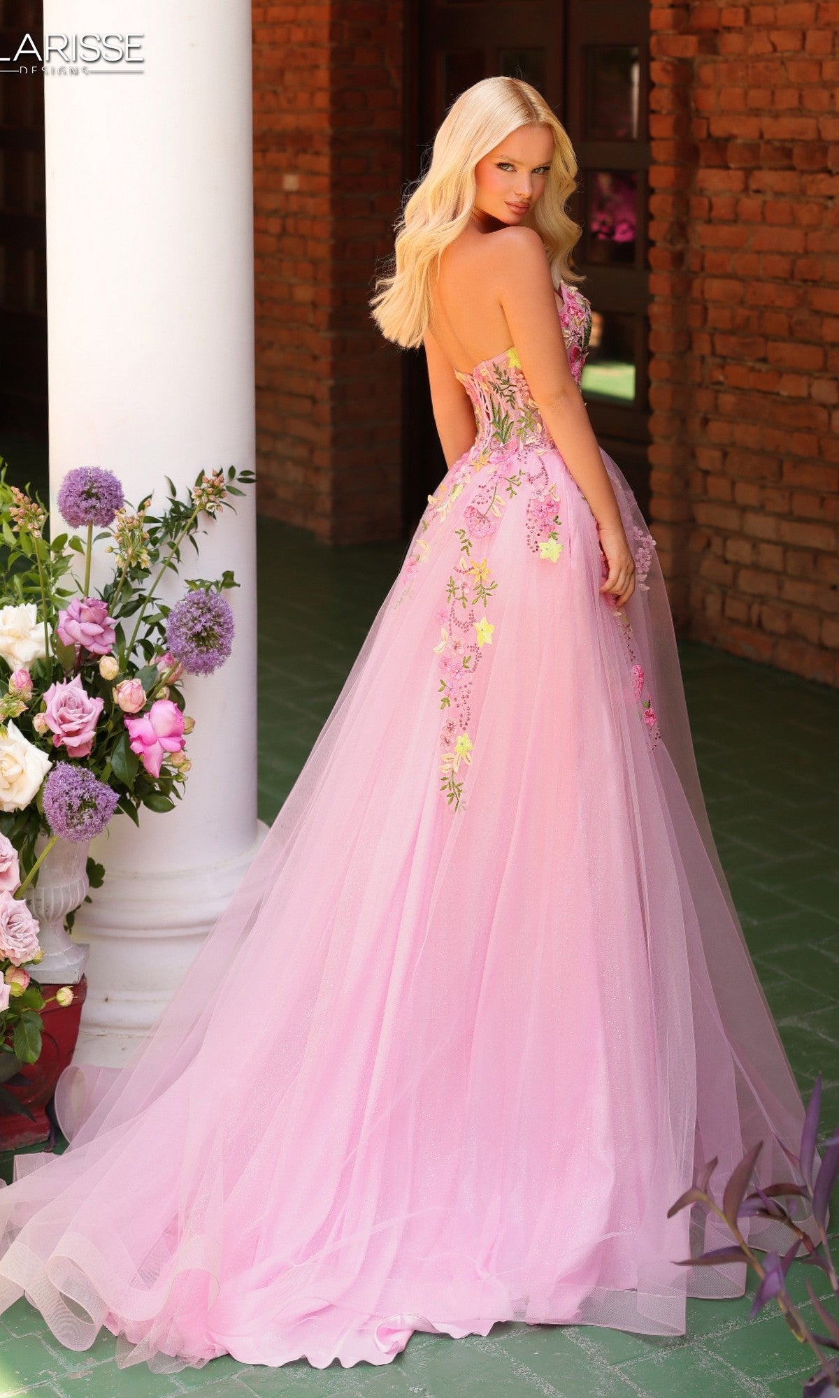 A woman wearing the Long Prom Dress: Clarisse Designs 62138, a strapless ball gown with floral lace embroidery and a sweetheart neckline, stands on a green-tiled floor near a brick wall and white column, looking over her shoulder.